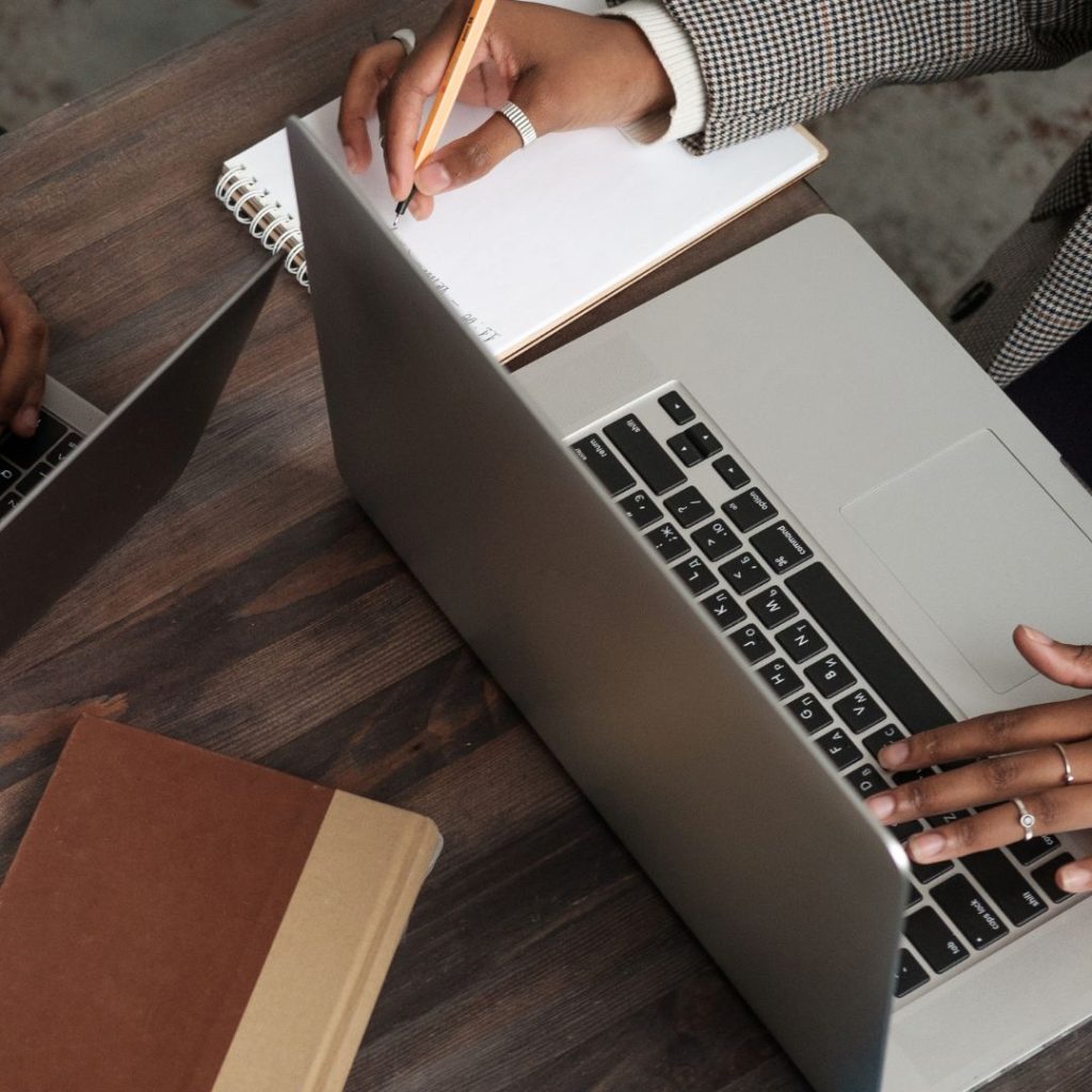 Two people working at a wooden table, one using a laptop and the other writing in a notebook. The scene conveys collaboration and productivity.